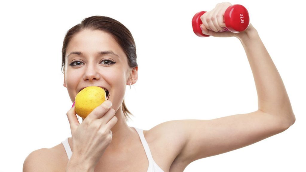 Mulher comendo fruta e segurando anilha em treino na academia.